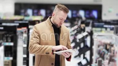 年轻英俊的长胡子男穿着棕色外套，在购物中心使用数字平板电脑。